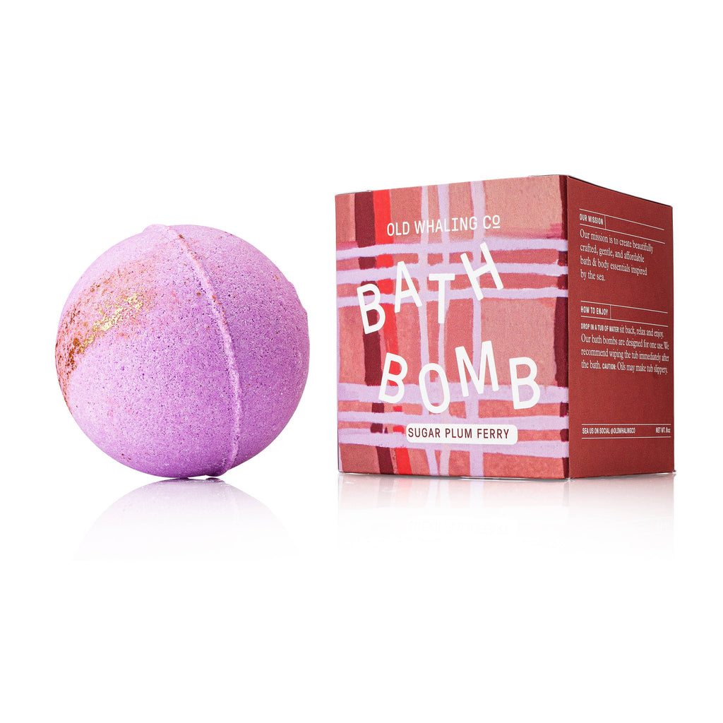 A spherical pink and gold bath bomb next to its pink and red plaid packaging.