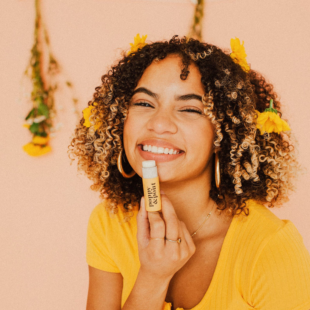 A person with yellow flowers in their hair holding a Lemon Bloom Lip Balm up to their lips.