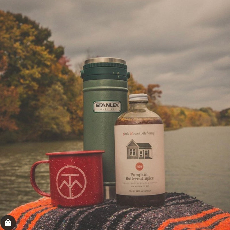 A grainy image of a camper mug, Stanley thermal cup, and Pumpkin Butternut Spice Syrup in front of autumnal foliage and a river.